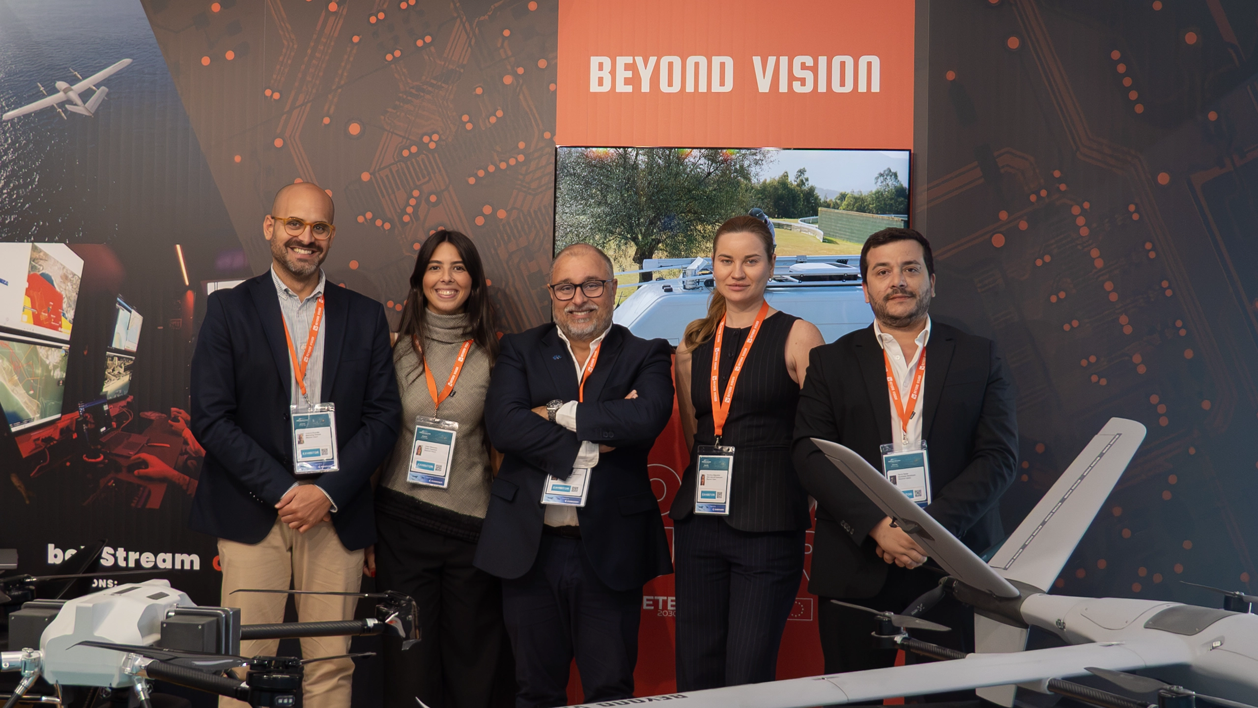 Five Beyond Vision team members at the company stand at Farnborough, with a VTOL fixed-wing drone and a quadcopter on display.
