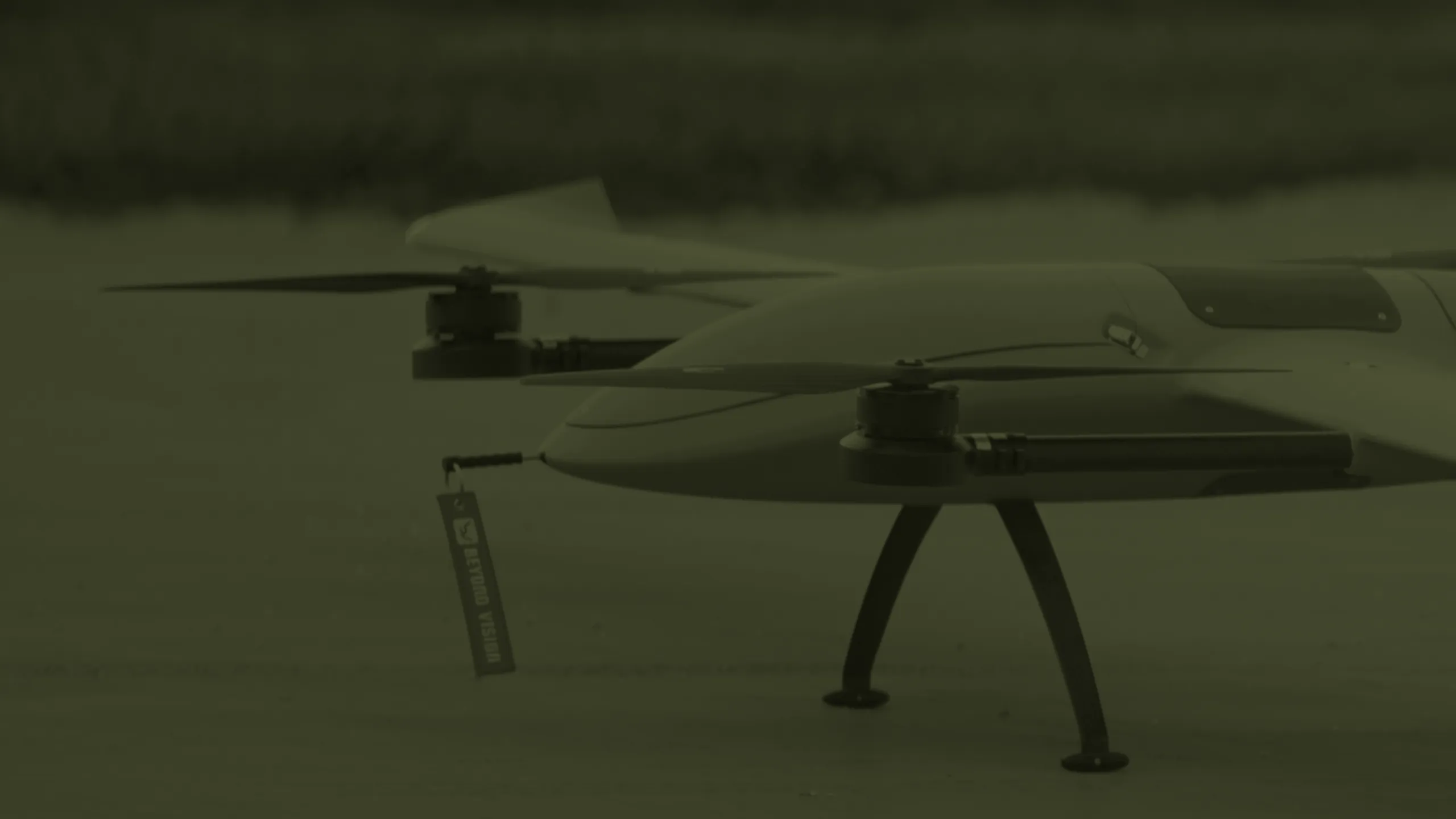 Nose of a Beyond Vision fixed-wing VTOL drone on the ground, front rotors at rest and a Beyond Vision tag hanging from the nose