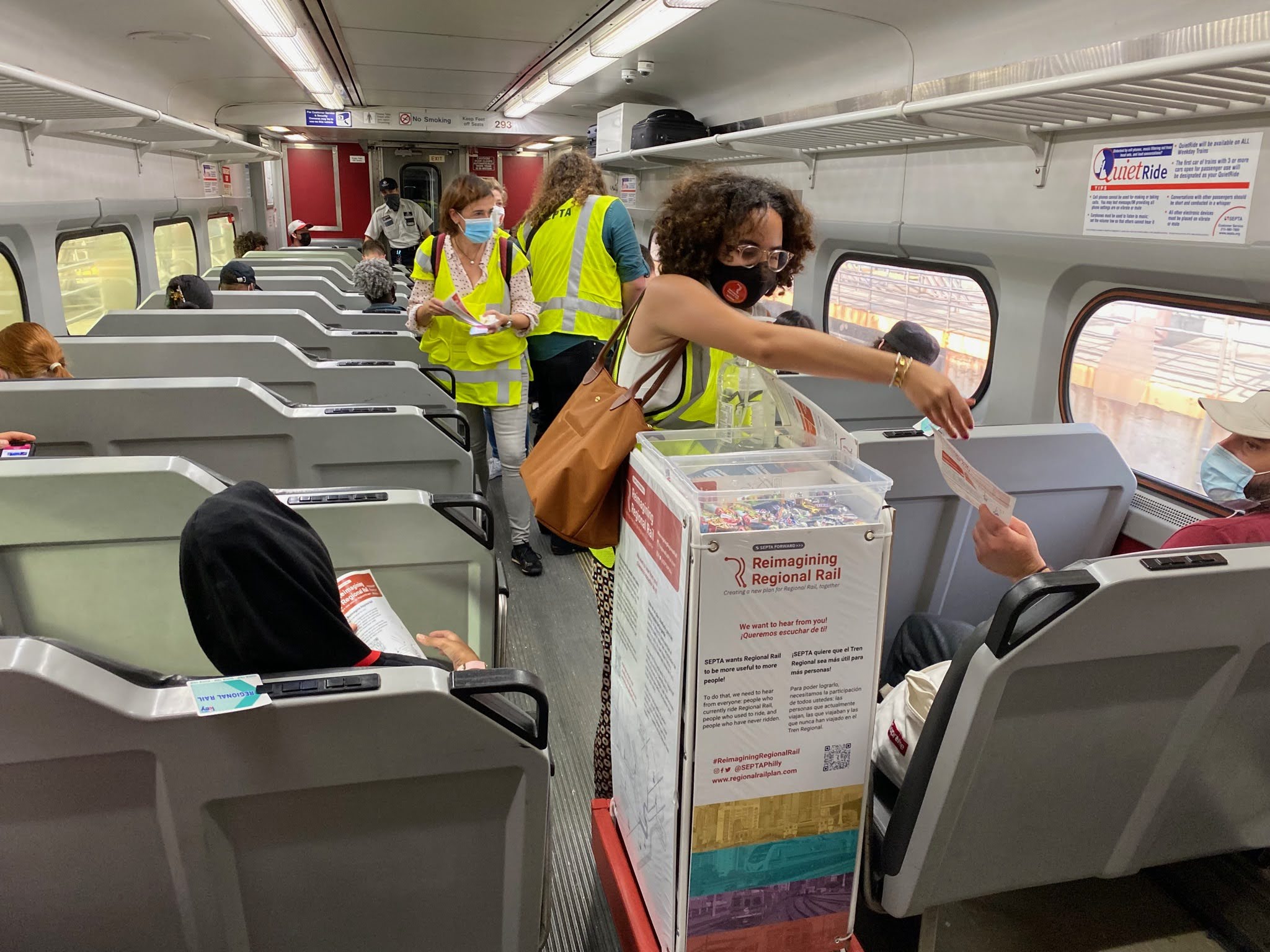 A woman inside a regional rail train car hands out snacks and a survey to passengers.