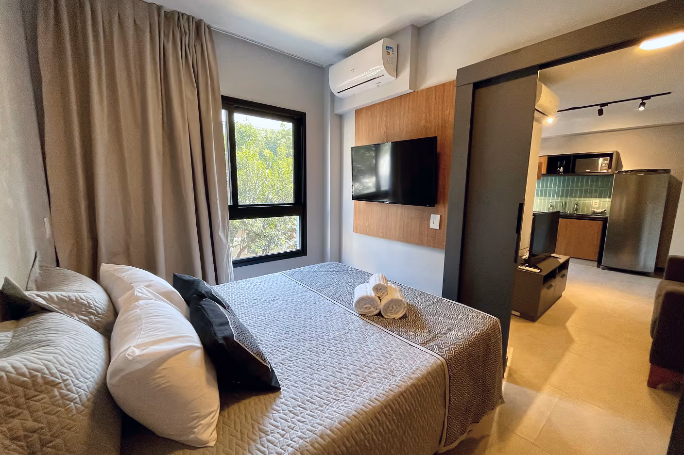 Quarto de studio Indepy com cama de casal, TV em painel de madeira e cozinha integrada ao fundo, formato voltado para locação short stay em São Paulo.