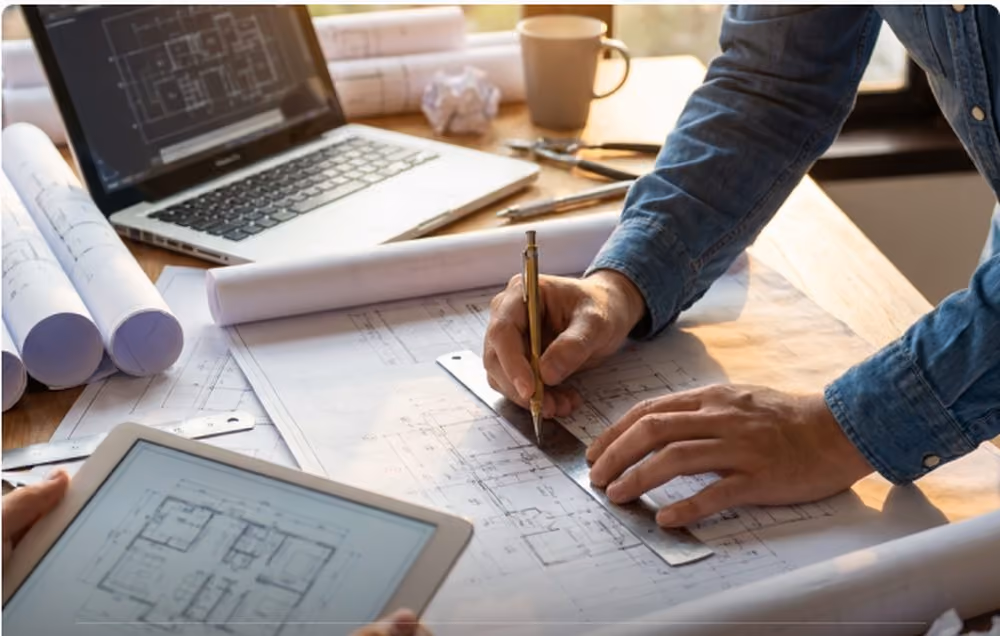 Mãos desenhando sobre planta arquitetônica em prancheta, com laptop e tablet exibindo projetos — fase de projeto de um empreendimento Indepy.