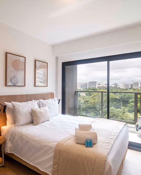 Quarto de apartamento Indepy com cama de casal, quadros na parede e porta de vidro para a varanda com vista para área verde, em São Paulo.