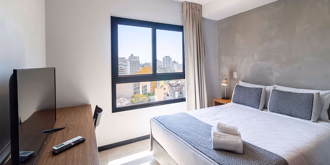 Quarto de studio Indepy com cama de casal, parede em cimento queimado e janela com vista para os prédios de São Paulo, formato short stay.