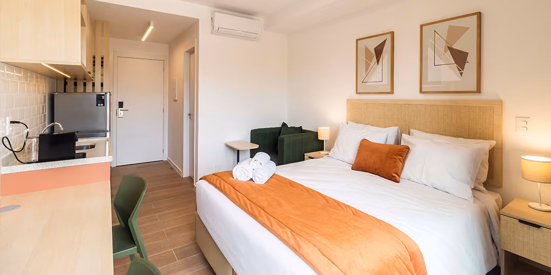Studio Indepy mobiliado com cama de casal, roupa de cama em tons de laranja, quadros na parede e cozinha compacta ao fundo, pronto para locação por temporada.