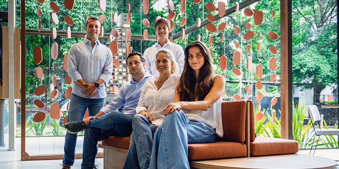 Equipe da Indepy reunida no lounge da sede, com sofá de couro caramelo e painel de peças suspensas diante de janelas com vista para a vegetação, em São Paulo.