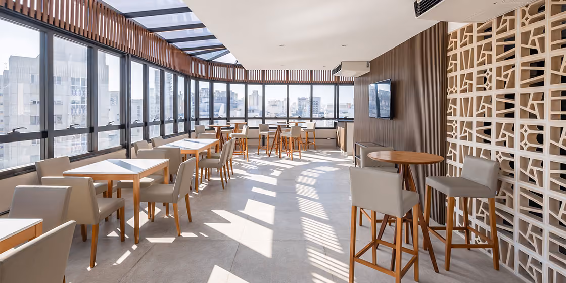 Espaço de coworking e convivência de empreendimento Indepy, com janelas panorâmicas, mesas de madeira e painel vazado decorativo, área comum em São Paulo.