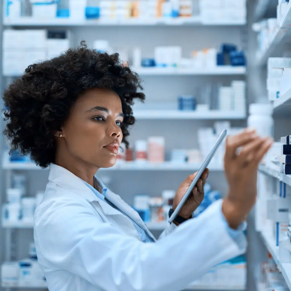 Pharmacist in white coat holding tablet, selecting medicine from pharmacy shelf.