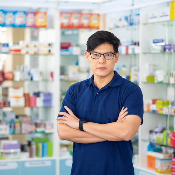 Man wearing glasses and a navy polo stands with arms crossed in a pharmacy with shelves of medicine.