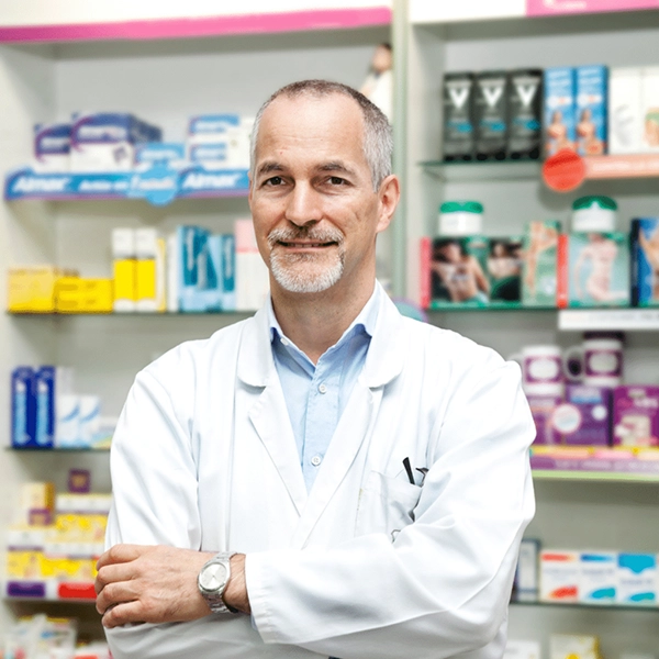 Middle-aged pharmacist in white coat standing with arms crossed in front of shelves with medicines.