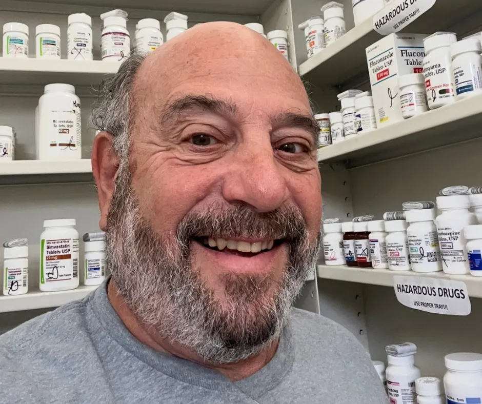 Smiling man with beard standing in front of shelves filled with prescription medication bottles.