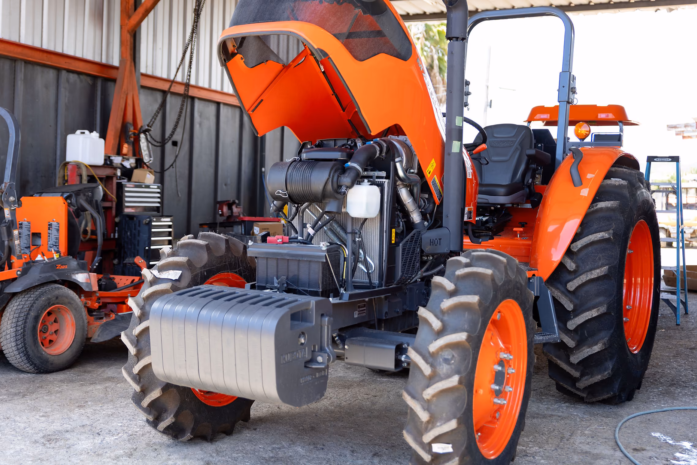 Tractor agrícola naranja con el capó abierto mostrando el motor, estacionado en un taller con herramientas y otro tractor al fondo.