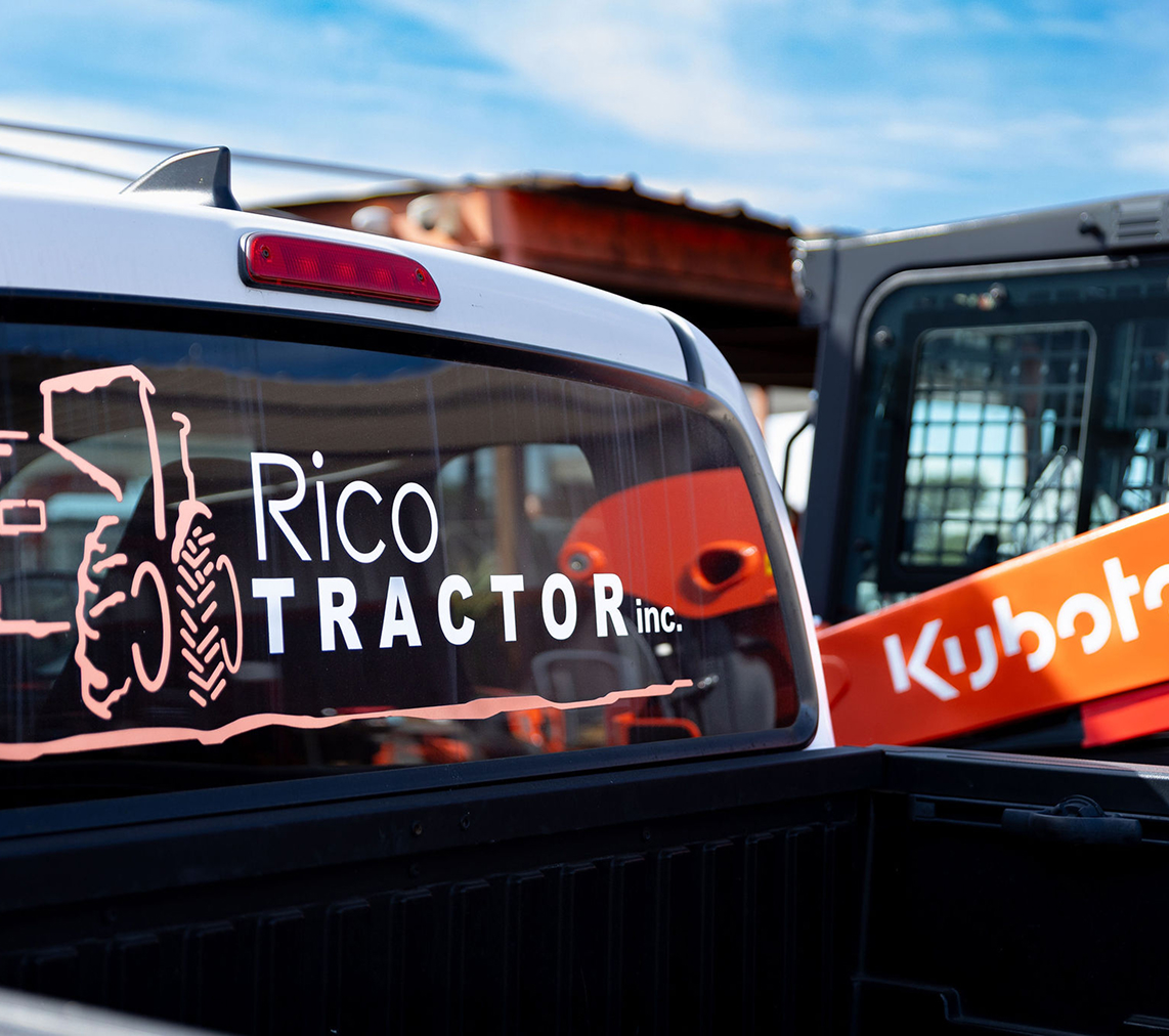 Ventana trasera de una camioneta con el logo de Rico Tractor Inc. y un dibujo de un tractor, con maquinaria Kubota naranja visible detrás.