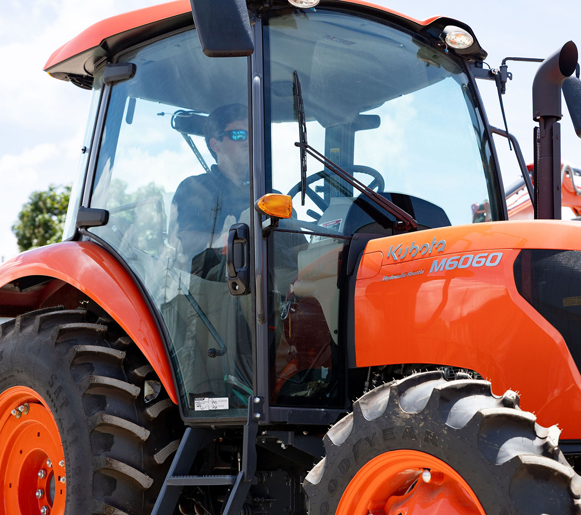 Hombre con gafas de sol sentado dentro de un tractor Kubota naranja modelo M6060, visible desde una ventana frontal de vidrio.