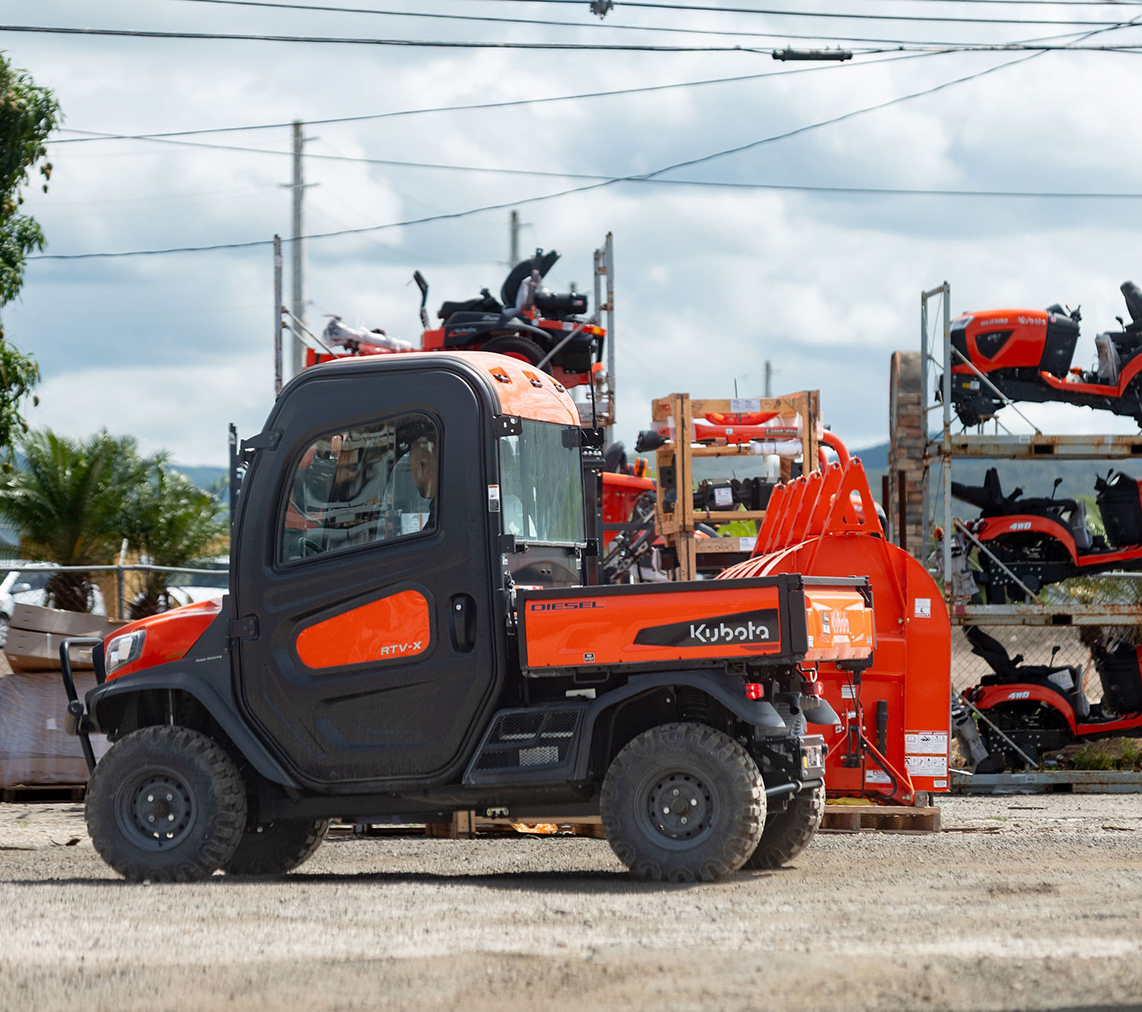 Vehículo utilitario Kubota RTV-X de color negro y naranja estacionado en un área con equipo agrícola y maquinaria apilada al fondo, bajo un cielo nublado.