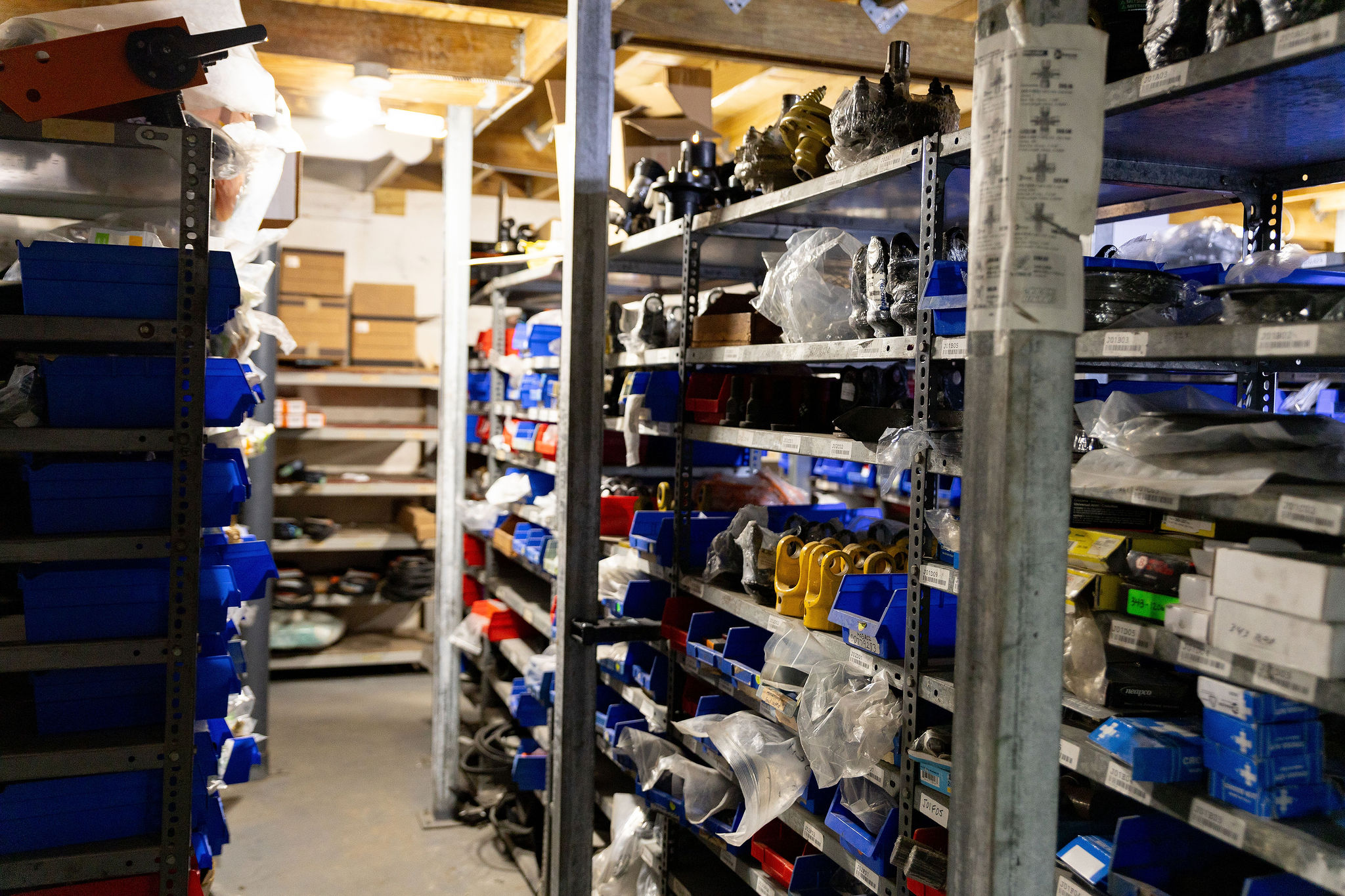 Pasillo de un almacén con estantes metálicos llenos de cajas azules y varias piezas mecánicas organizadas en bolsas plásticas y compartimentos.