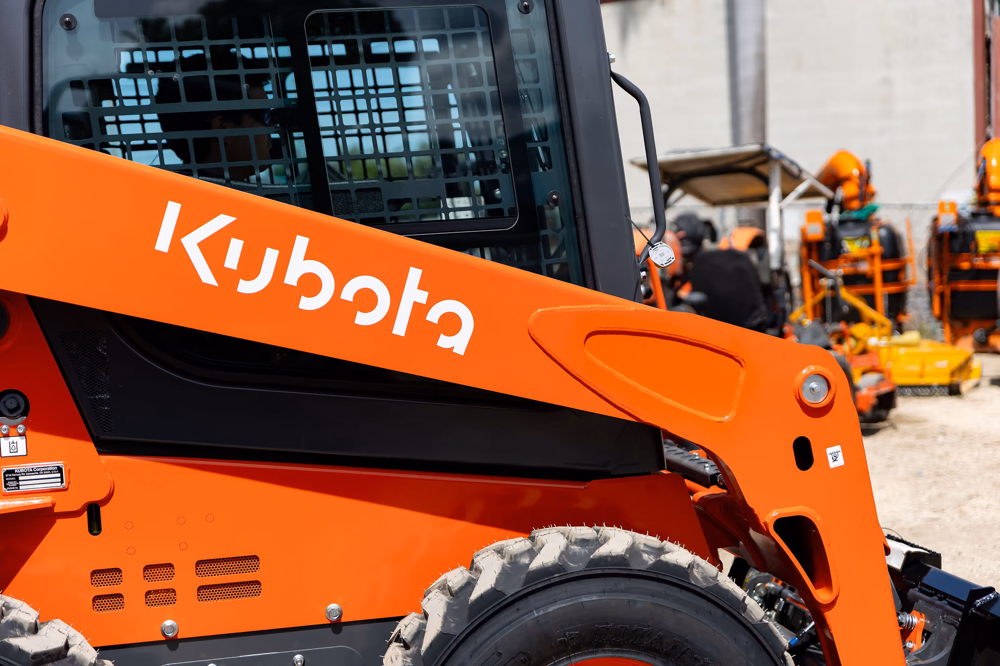 Maquinaria de construcción Kubota de color naranja con una rueda grande visible y estructura metálica negra en un patio con más equipos naranjas al fondo.