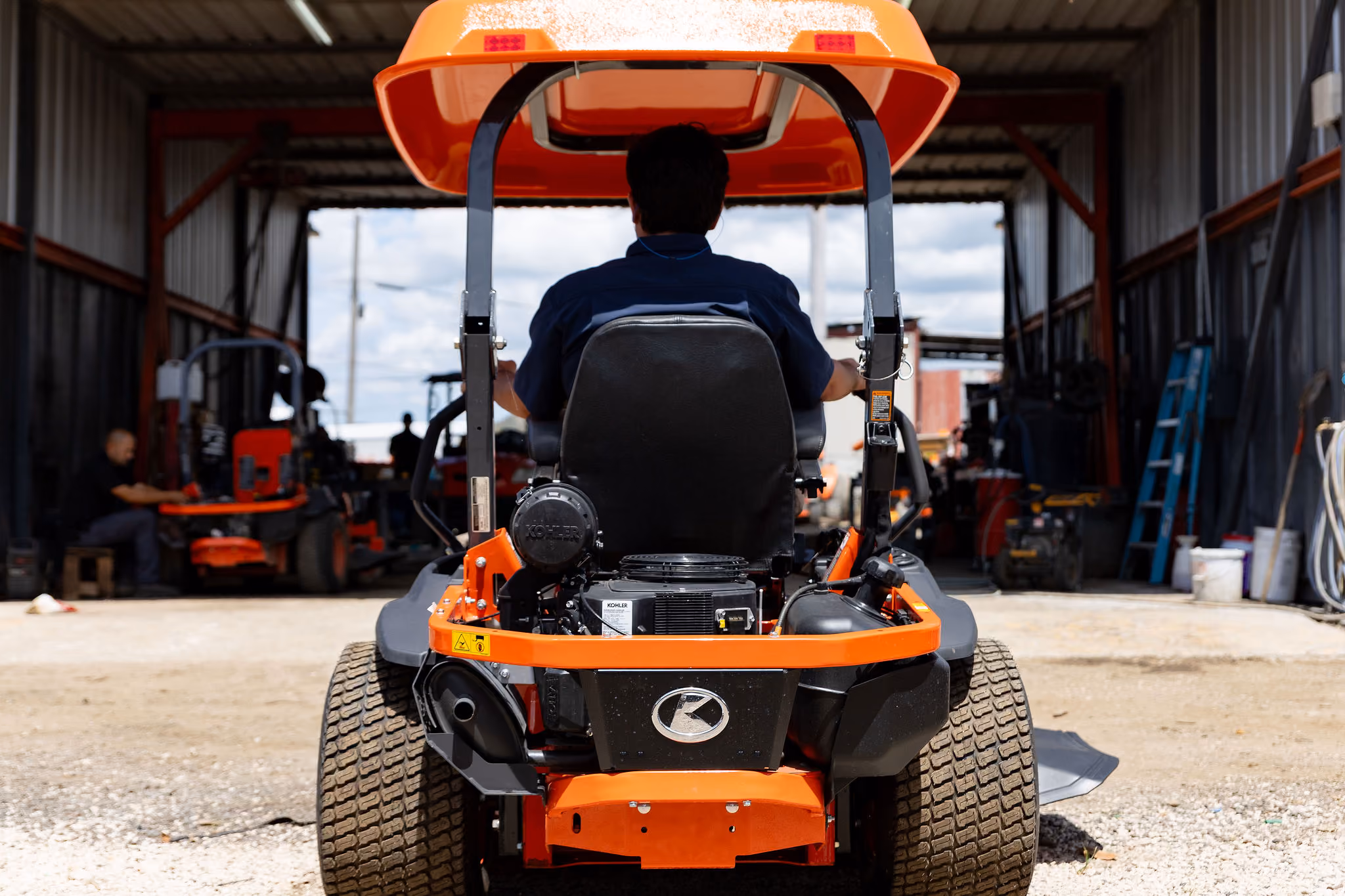 Persona sentada en un tractor naranja visto desde atrás en un taller con otras máquinas y herramientas.
