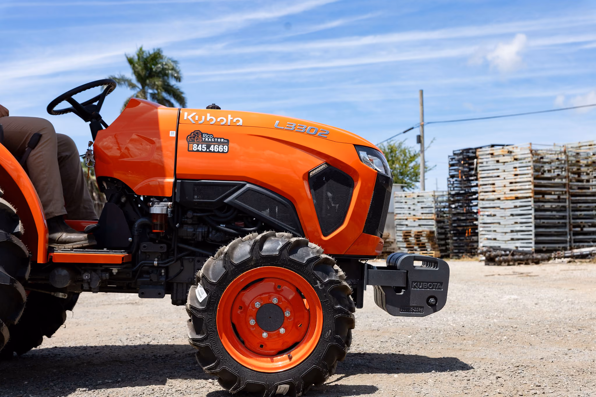 Tractor Kubota L3302 de color naranja con un neumático delantero grande, en un área al aire libre con montones de paletas de madera apiladas y un cielo azul con nubes.