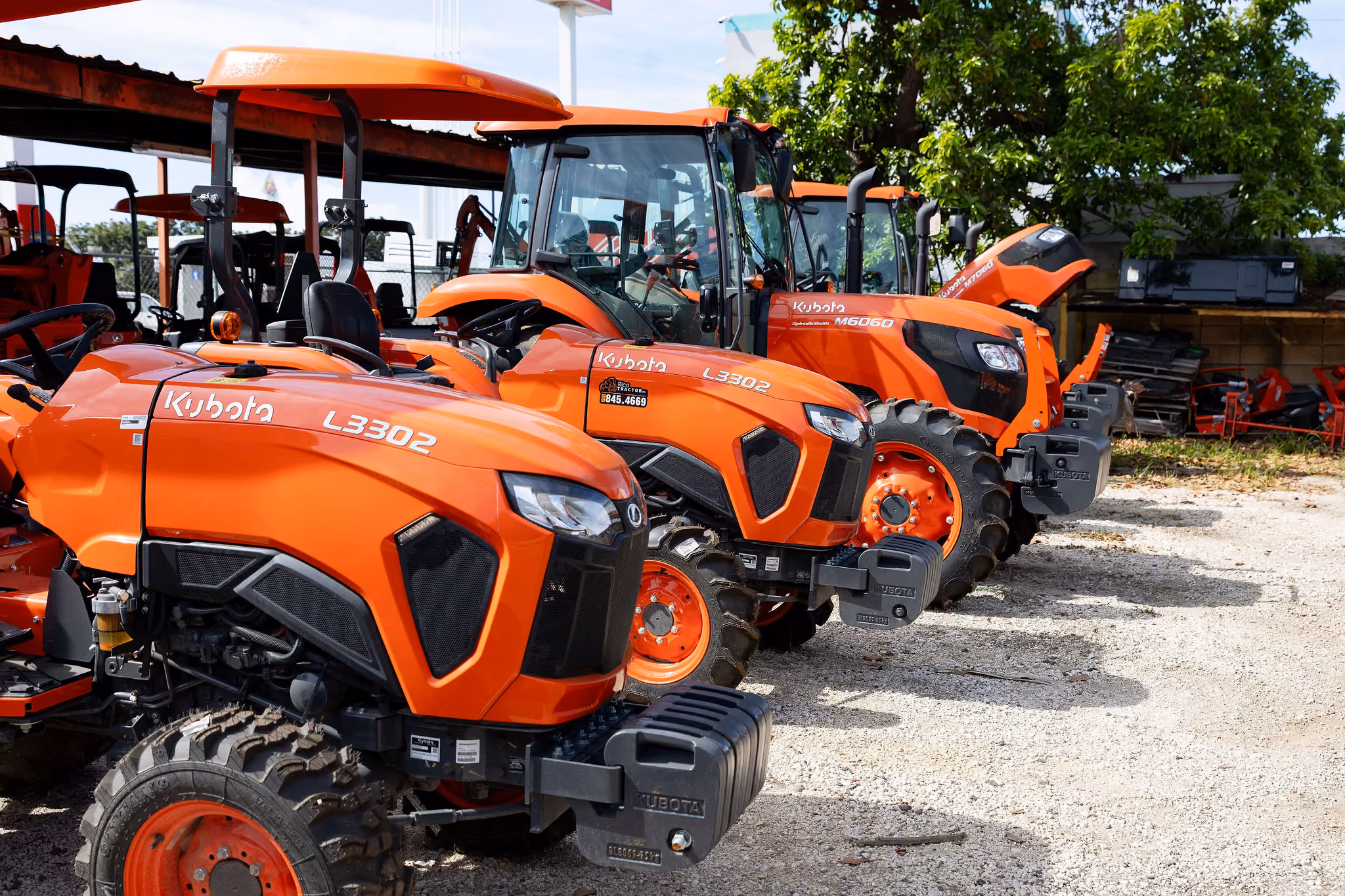 Varias tractores agrícolas de color naranja Kubota estacionados en un terreno con grava bajo un cielo parcialmente nublado y árboles verdes al fondo.