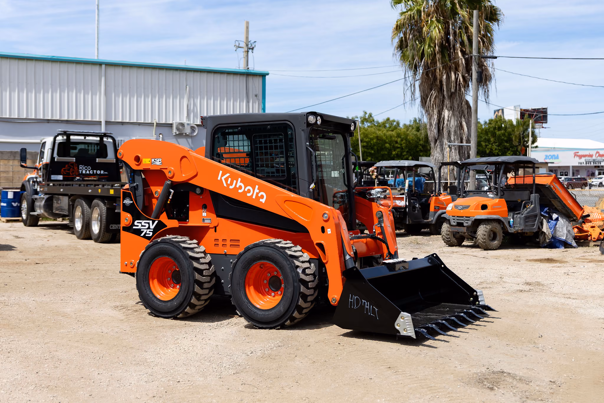 Minicargadora naranja Kubota SSV75 estacionada en un área de trabajo con tierra, con varios vehículos y maquinaria detrás y una palmera a la derecha.