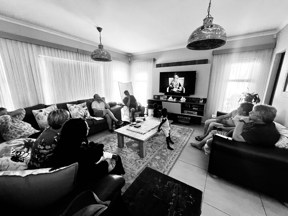 Black and white photo of a living room with eight people sitting on sofas around a low rectangular table, two children standing in front of a TV screen displaying a person, and large pendant lights hanging from the ceiling.