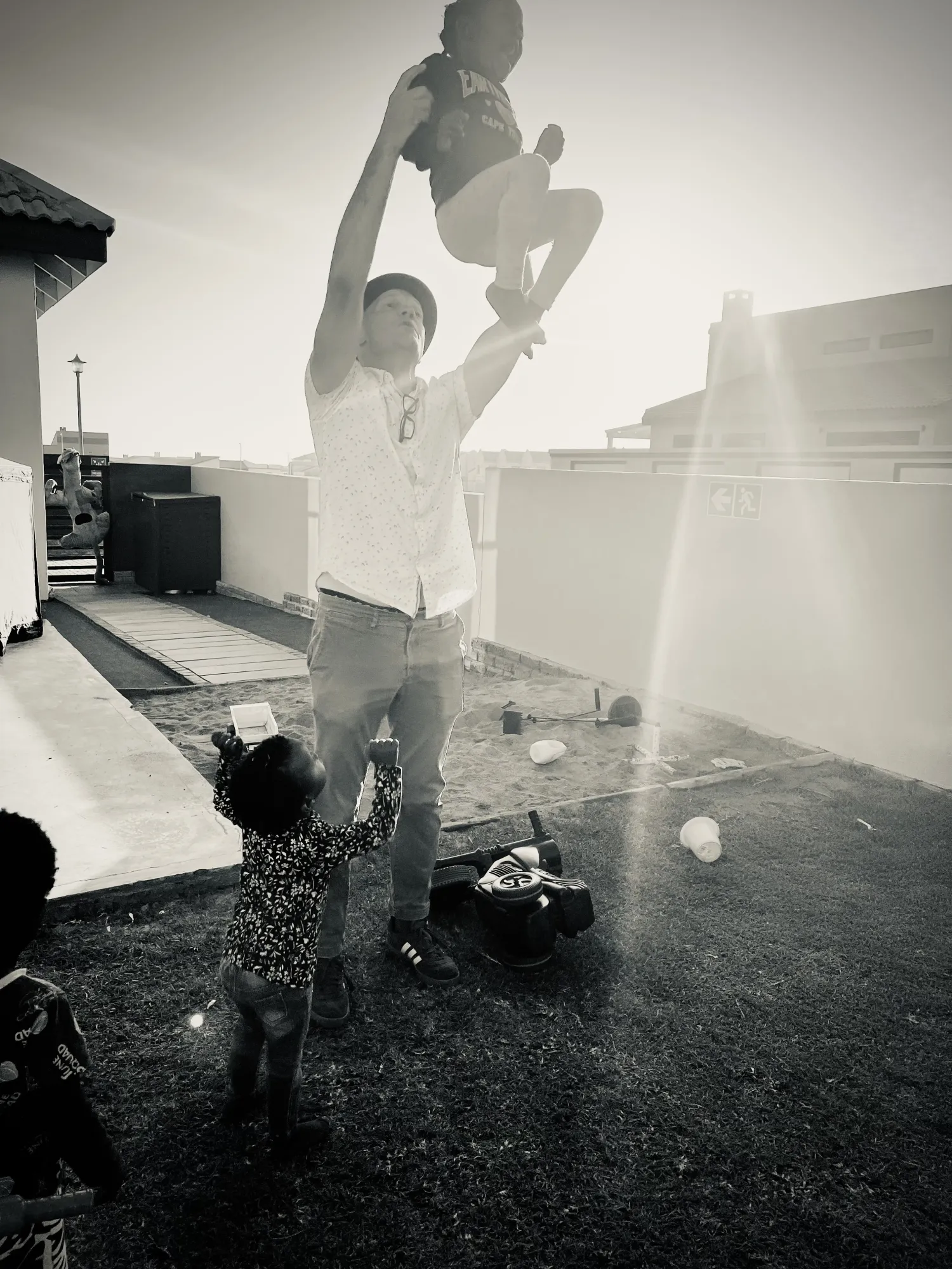 A man lifting a laughing child overhead outdoors while a toddler holds onto his leg