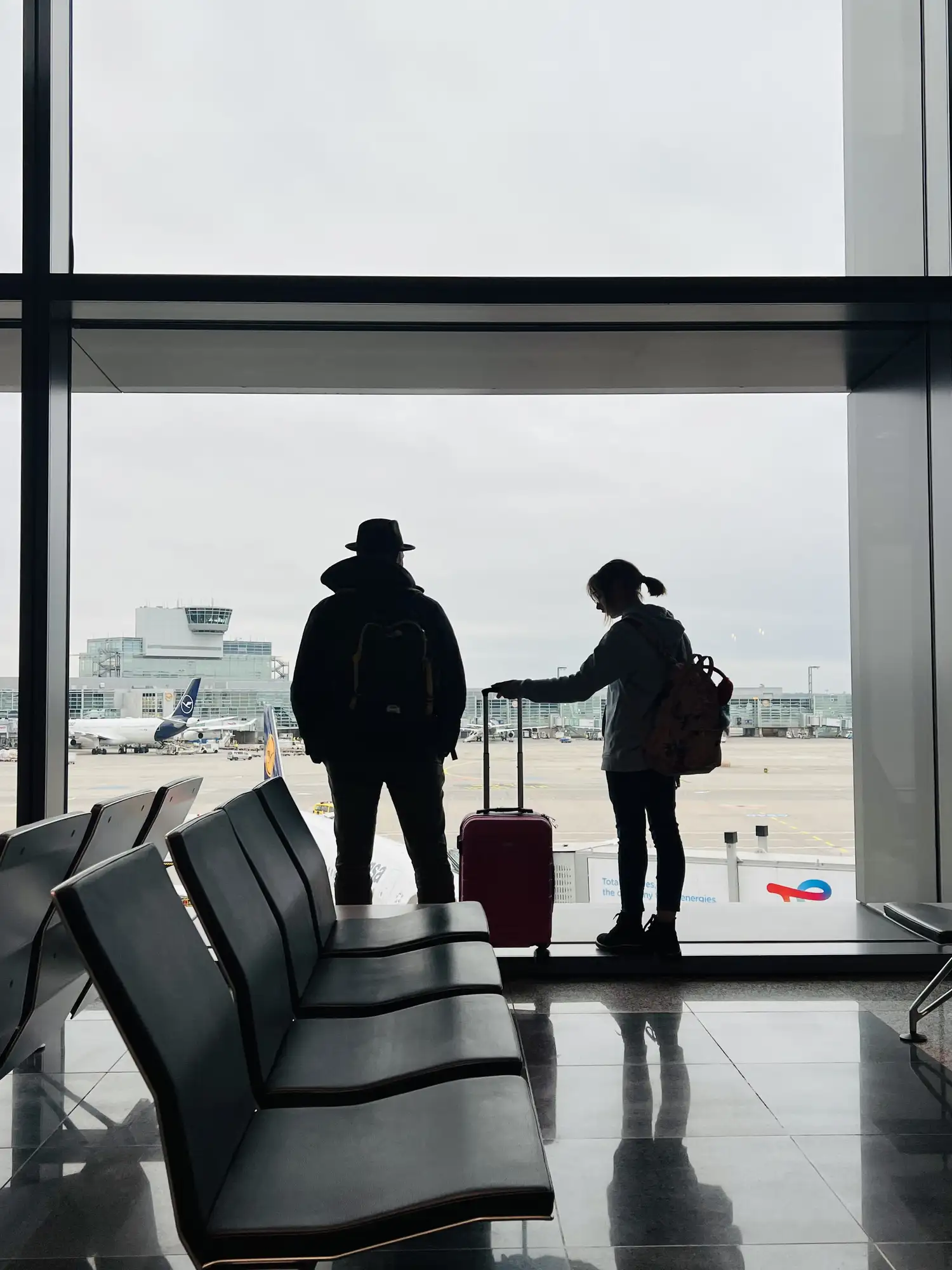 Two filmmakers with luggage watching planes from an airport window before traveling to a film location