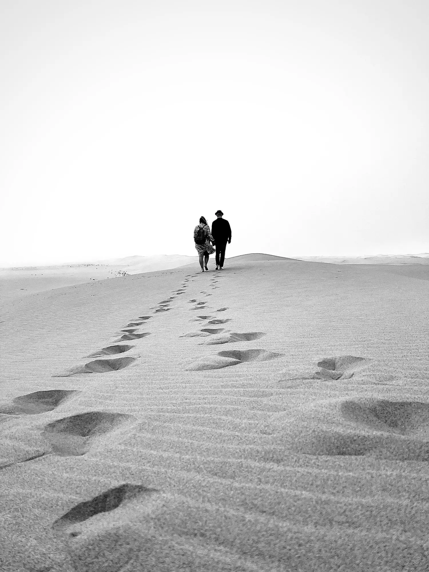 A couple walking across desert sand dunes, leaving a trail of footprints behind them