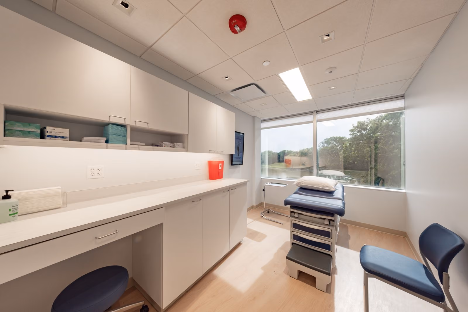 Bright medical exam room with cabinets, a blue exam table, chair, and large window overlooking trees.