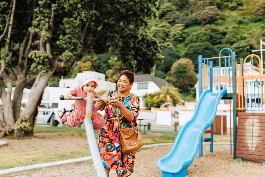 Child in pink dress and hat plays on seesaw with smiling adult in colorful dress at playground.