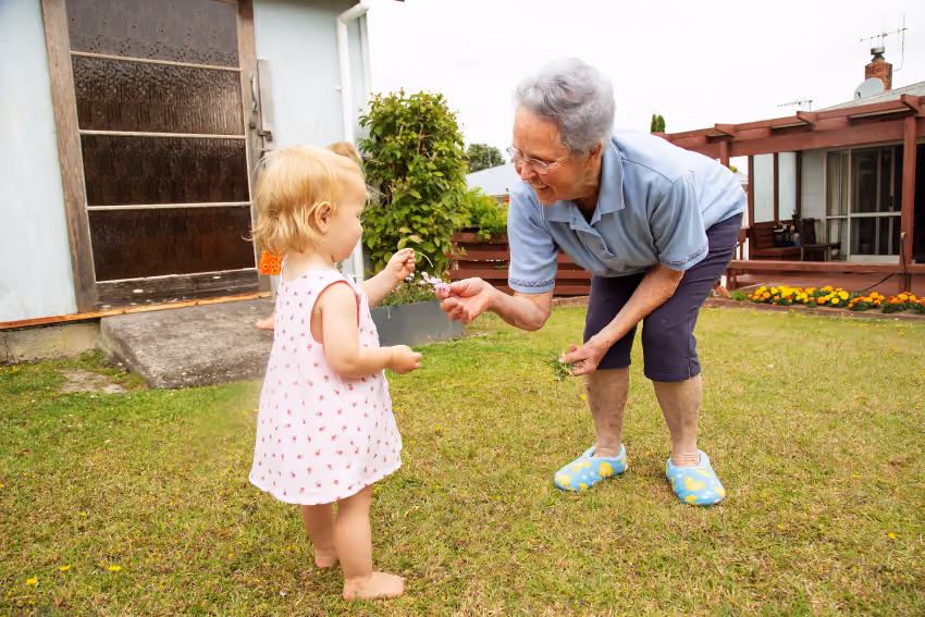 Elderly woman giving flowers to a toddler on grass outside a house.