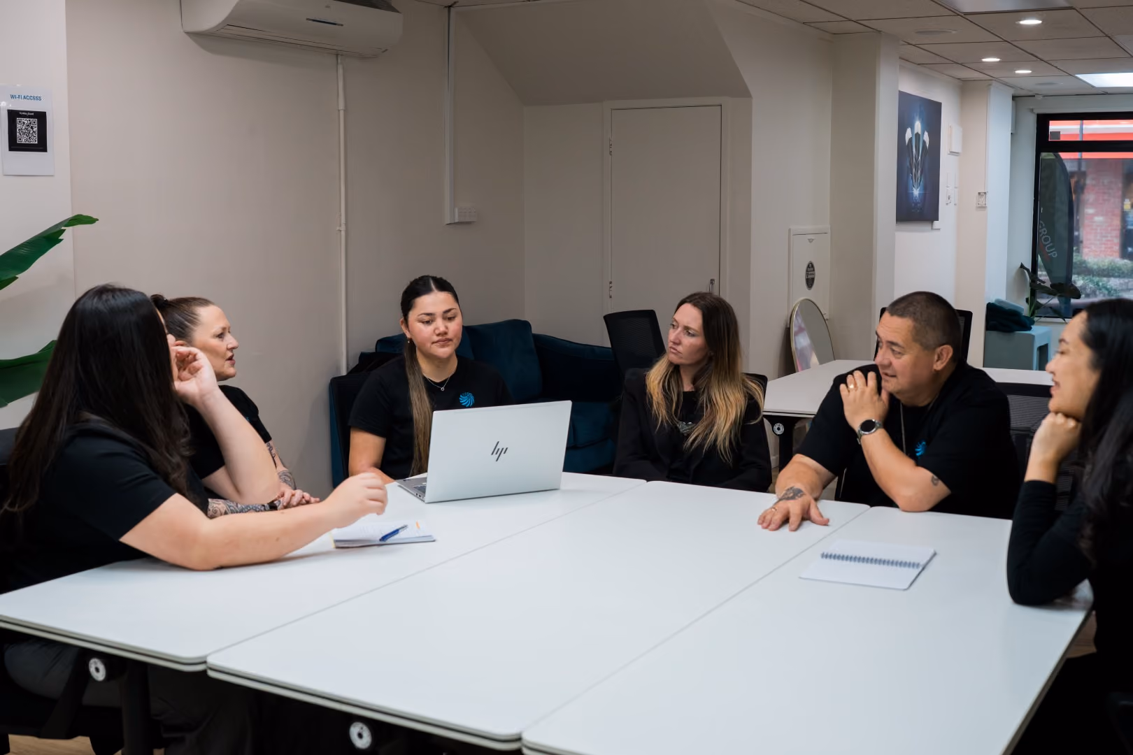 Three people gathered around a table looking at a laptop in a modern indoor space with plants.
