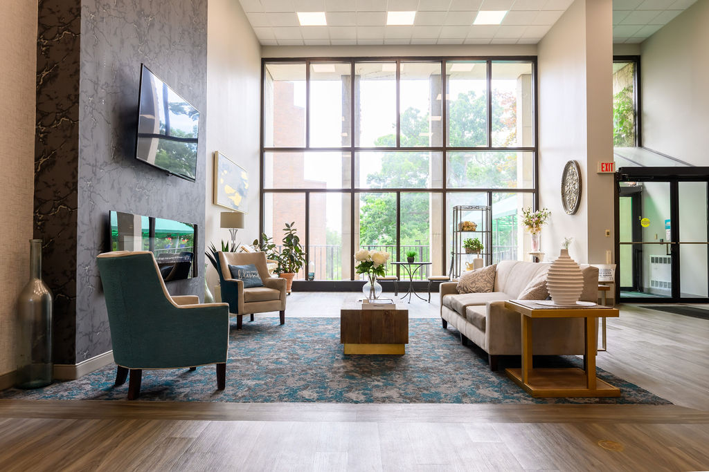 Bright living room with large windows, beige sofa, blue armchair, coffee table, and decorative plants.