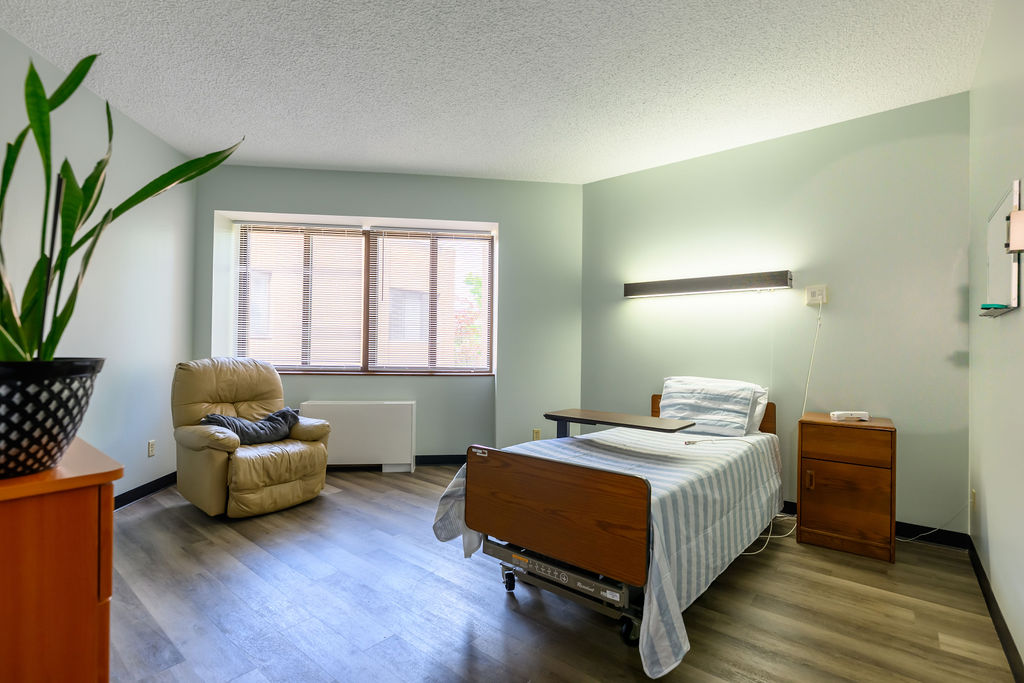 Simple room with a single hospital bed, beige recliner chair, wooden side table, and large window.