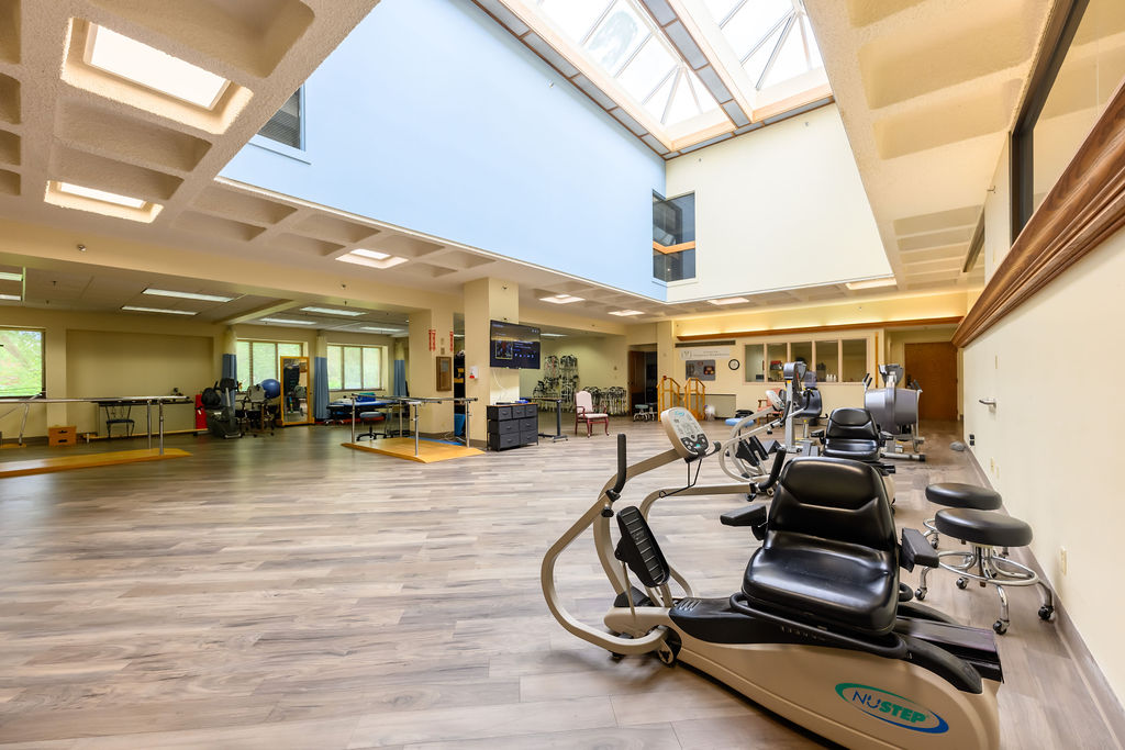 Bright rehabilitation gym with exercise machines, parallel bars, therapy mats, and skylights.