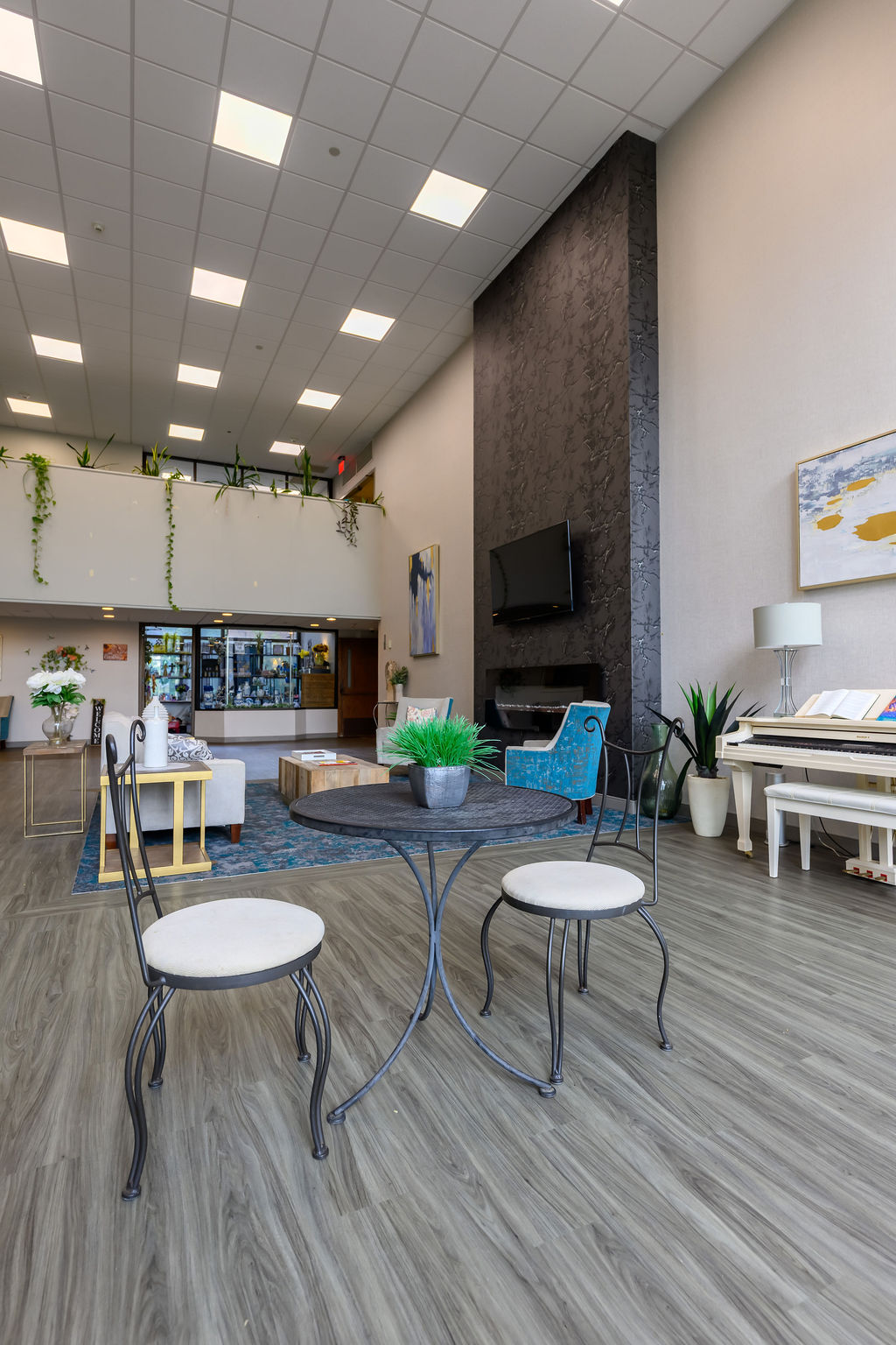 Modern lobby with two iron chairs and round table, blue armchair, TV, piano, plants, and high ceiling.