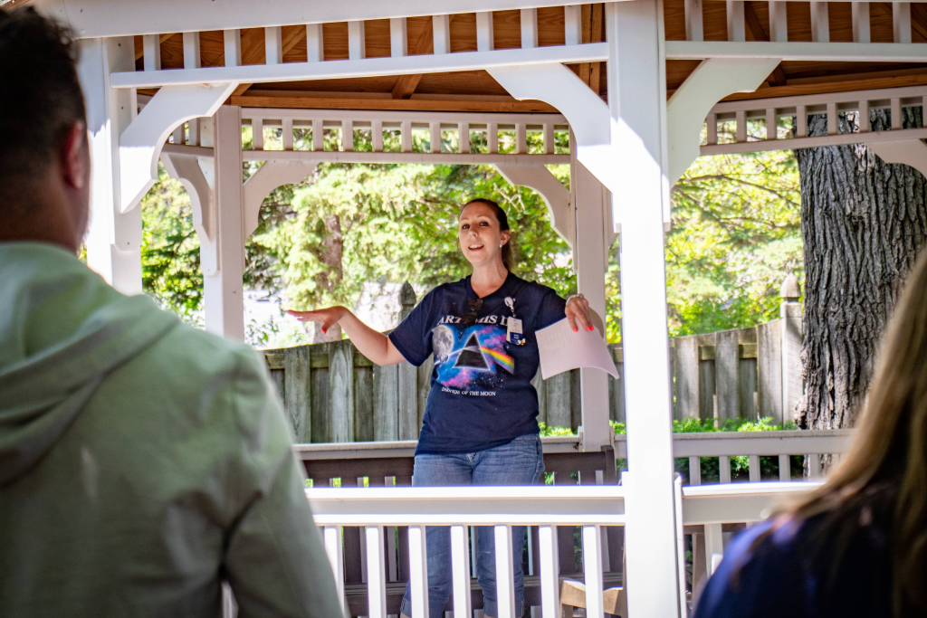 Woman in a navy shirt speaking and gesturing to people under a wooden gazebo outdoors.