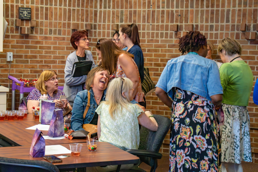 Group of women chatting and laughing around a table with drinks and gift bags in a brick-walled room.