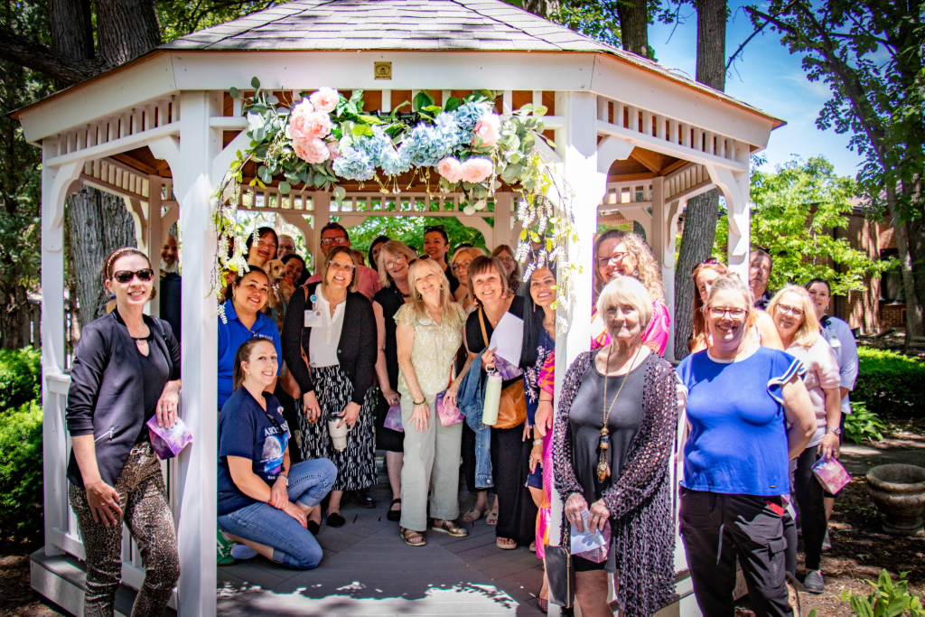 Group of smiling people posing inside and around a white gazebo decorated with pastel flowers.