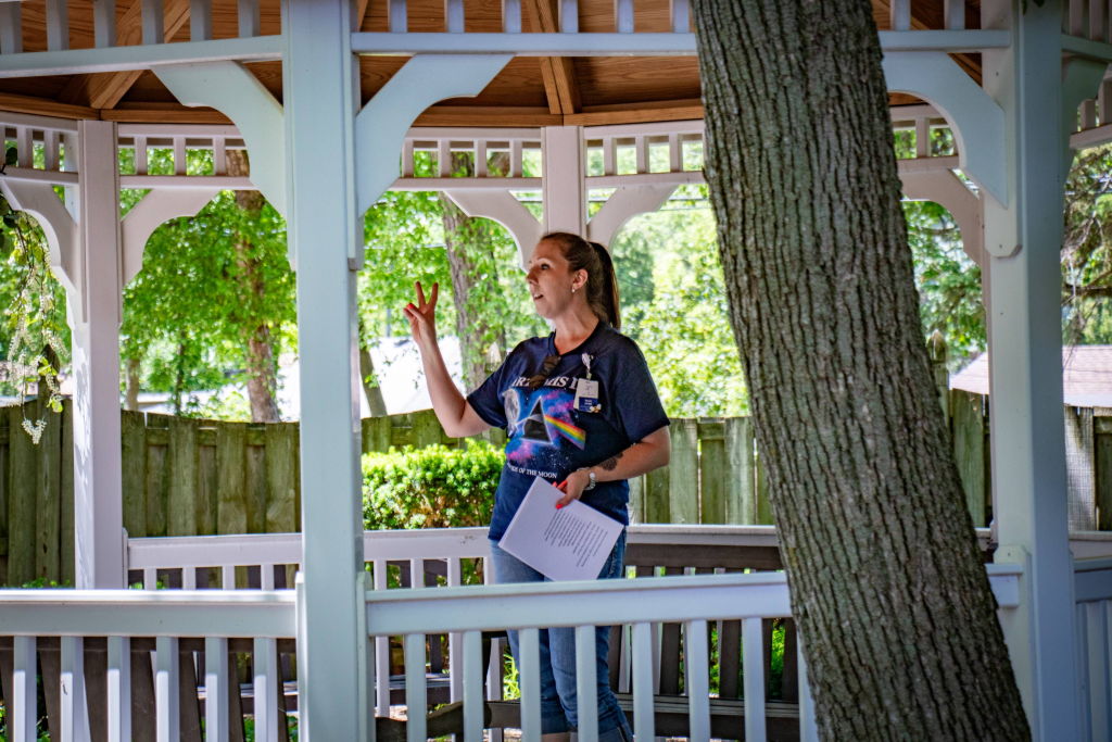 Woman in a gazebo holding papers and gesturing with her hand, surrounded by trees and wooden fence.