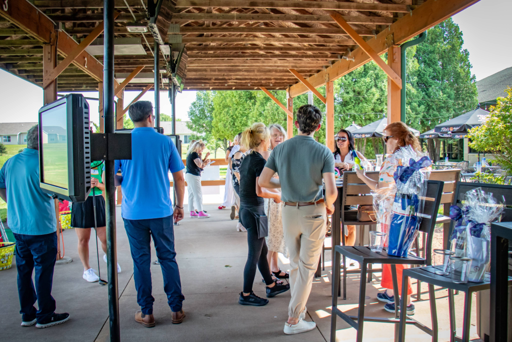 Group of people socializing under a wooden pavilion at an outdoor event with gift baskets on chairs.