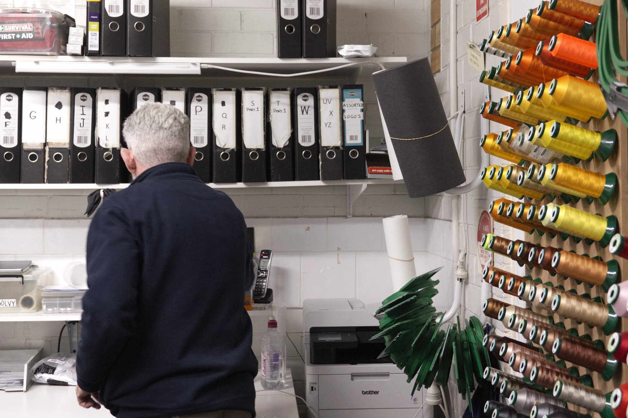 Uniforms Plus owner, Richard Innis, faces away while working on customer orders, he is surrounded by order folders and thread