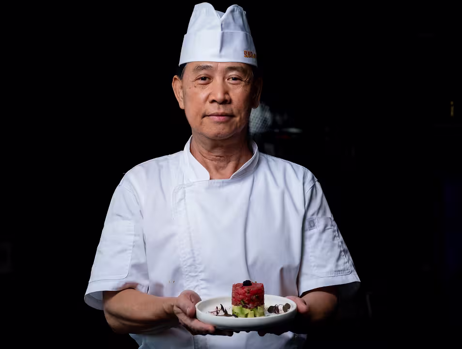 Chef in white uniform holding a plate with a layered dish of diced avocado and tuna garnished with caviar.