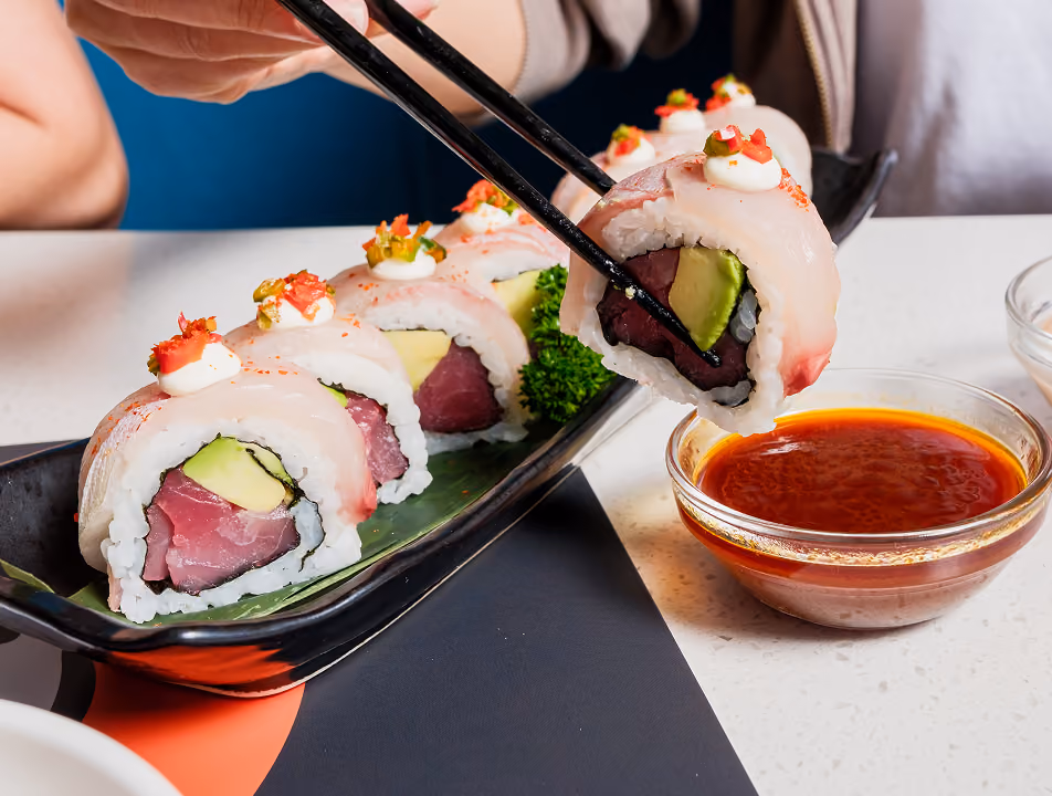 Chopsticks holding a piece of sushi roll with tuna, avocado, and white fish topped with a dollop of cream and diced peppers, near a bowl of soy sauce, with a black plate holding multiple sushi pieces garnished with green parsley on a white table.