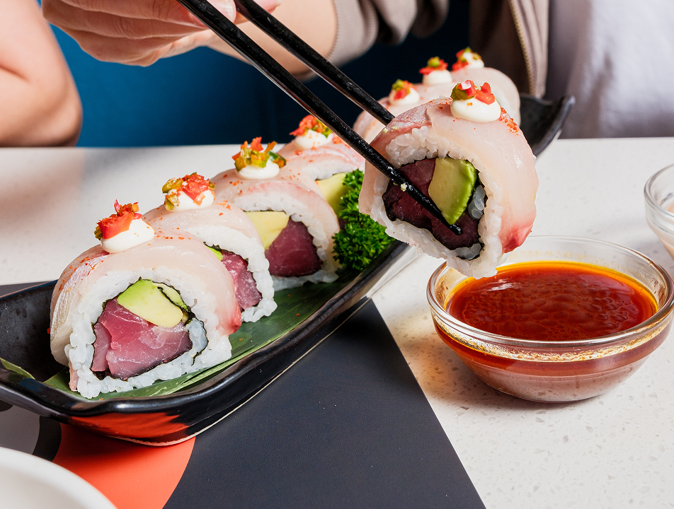 Chopsticks holding a piece of sushi roll with tuna, avocado, and white fish topped with a dollop of cream and diced peppers, near a bowl of soy sauce, with a black plate holding multiple sushi pieces garnished with green parsley on a white table.