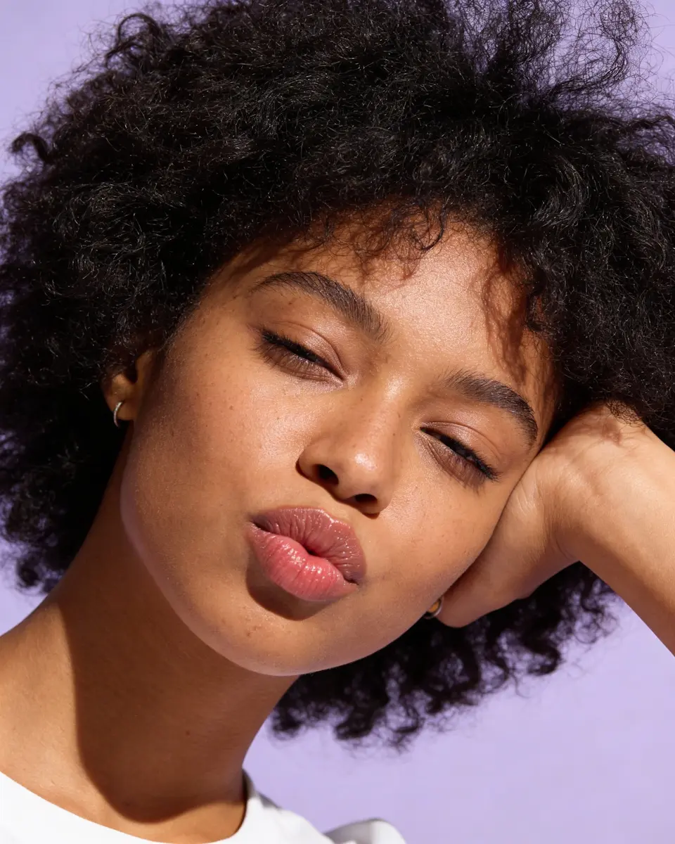 Close-up of a woman with curly hair puckering her lips against a lavender background.
