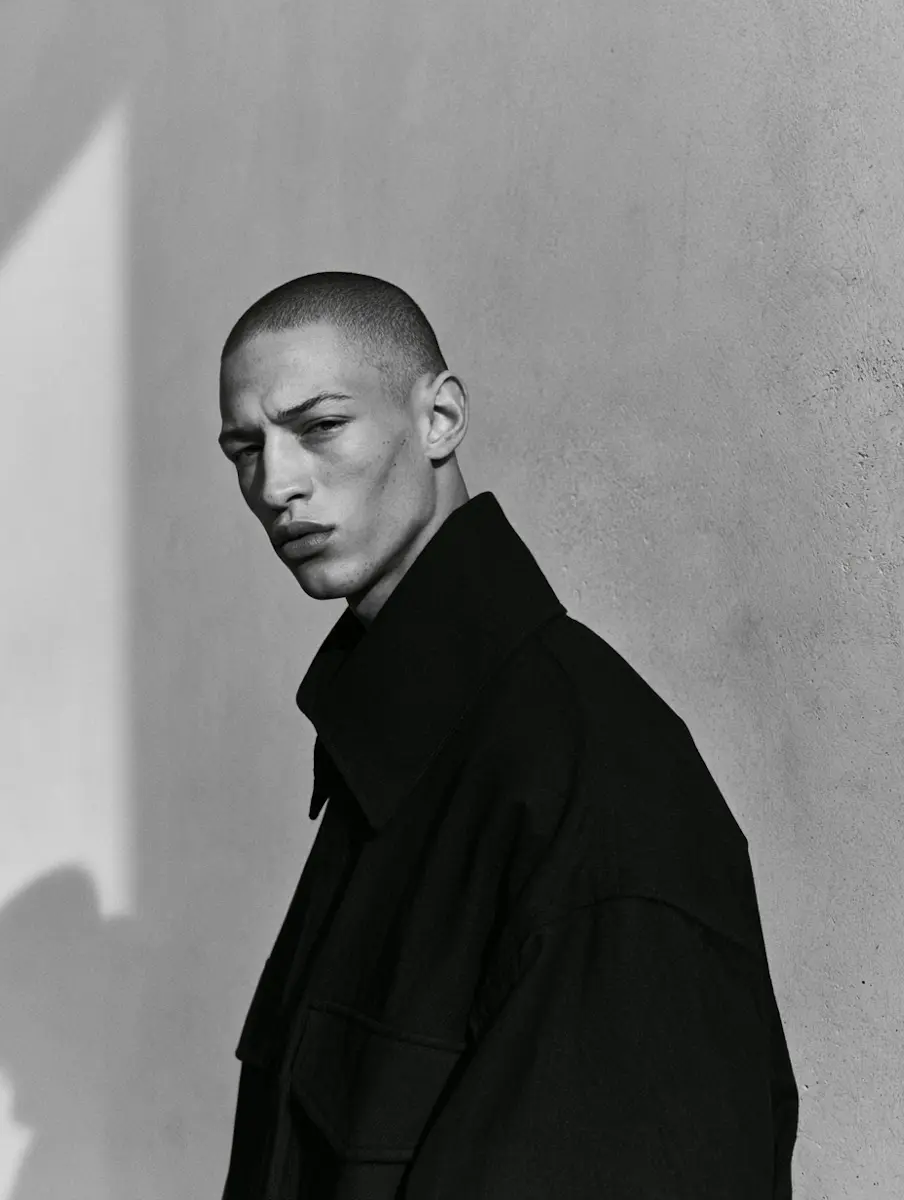 Black and white portrait of a man with a shaved head wearing a dark coat against a textured wall.