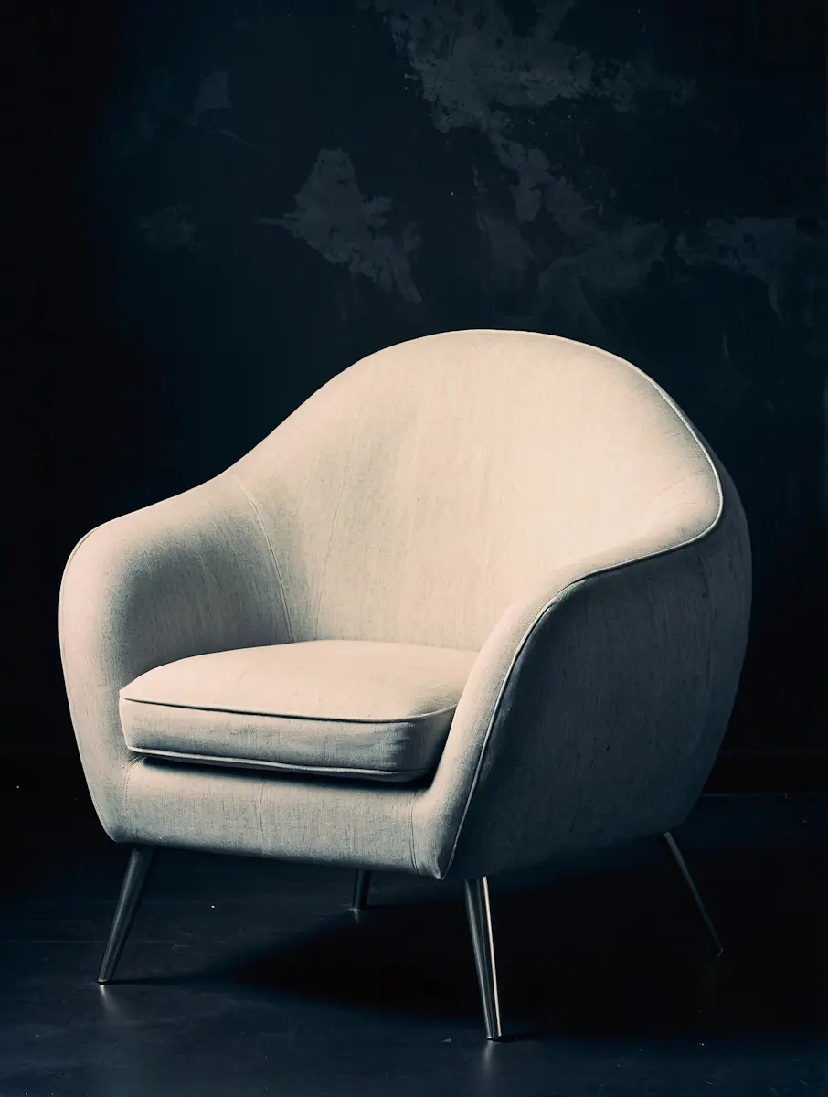 Modern beige armchair with rounded back and metal legs against a dark wall and floor.