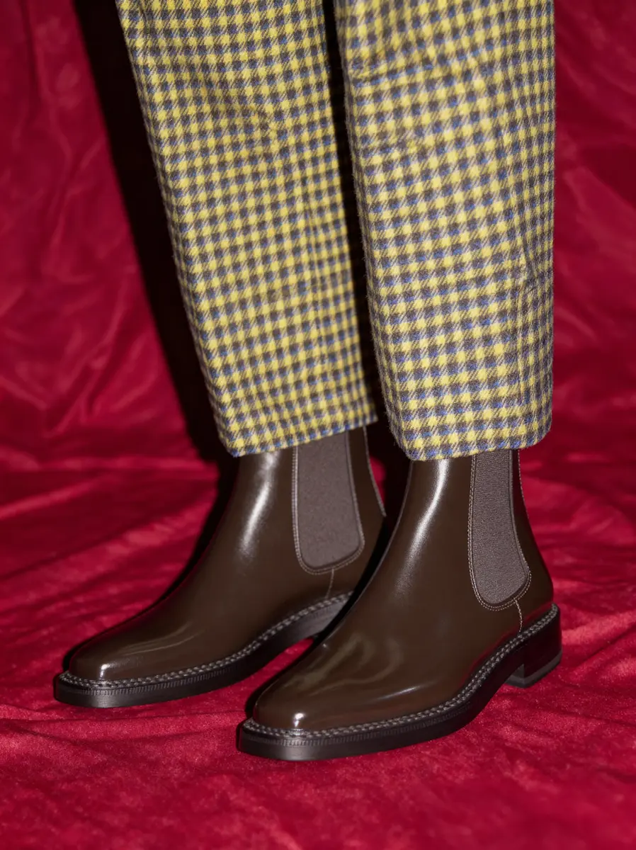 Pair of dark brown leather Chelsea boots worn with yellow and purple check pants on red fabric background.