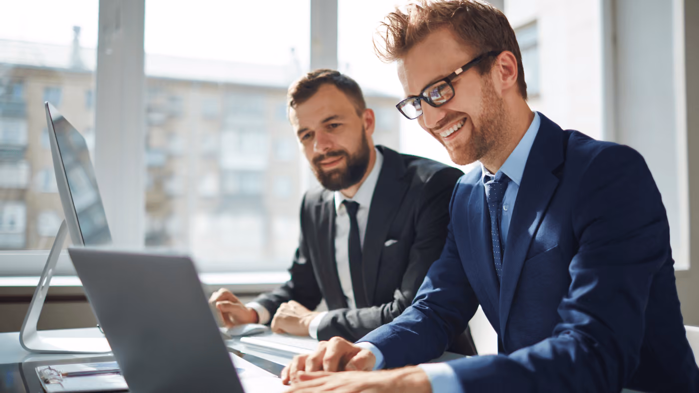 Two men wearing suits and glasses work side by side at a desk on their laptops